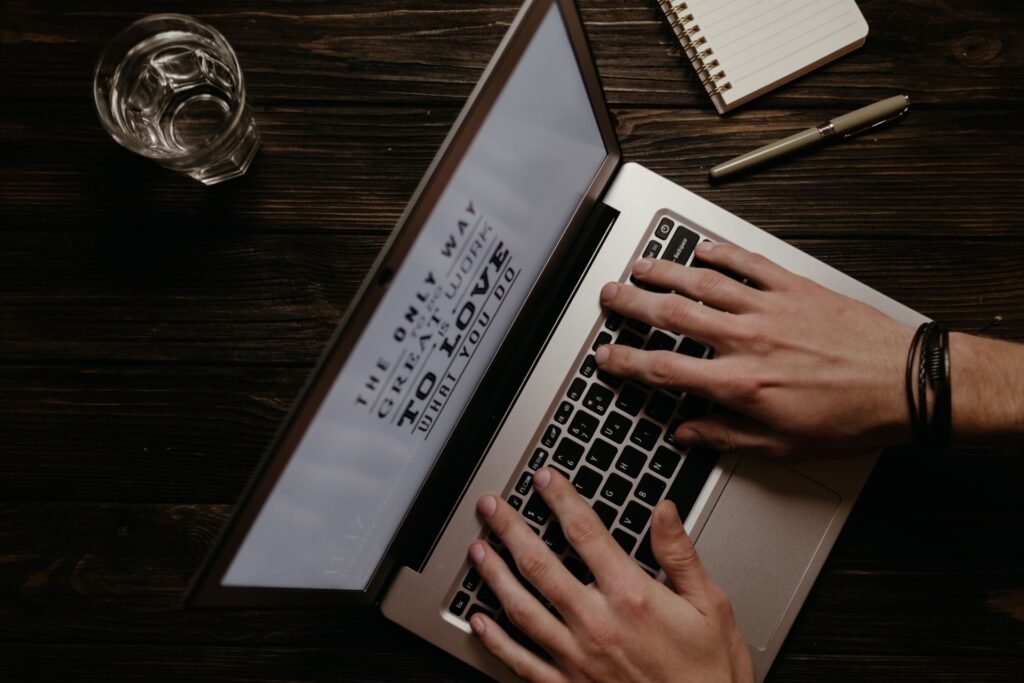 Mains qui tapent sur un ordinateur portable posé sur un bureau en bois sombre, carnet et verre d'eau
